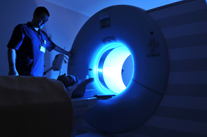 Picture of an MRI scanner. There is a medical worker (male) looking down at a male patient that is lying down about to go through the MRI scanner.