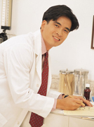 Picture of male Physician smiling and leaning over a counter top with a pen in his hand and paper under it. He is wearing a tie and medical coat.