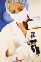 Picture of a female healthcare professional wearing a disposable medical head cap and a mask over her mouth. She is touching a microscope while wearing gloves.